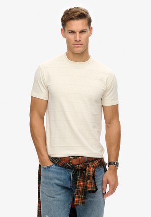 Beżowy t-shirt z krótkim rękawem w pionowe paski, okrągły dekolt i subtelne logo. Noszony z niebieskimi dżinsami i koszulą w kratę zawiązaną w talii.