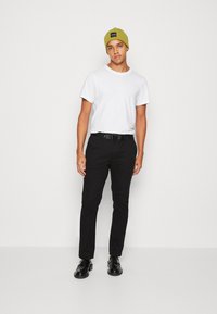 Pantaloni neri, maglietta bianca e berretto verde indossati con una cintura nera. Scarpe nere completano l'outfit, mostrando uno stile casual.