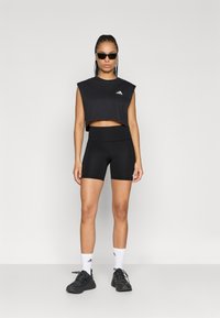 Crni kratki top bez rukava s Adidas logom, upareni s crnim šortsevima visokog struka. Model nosi crne tenisice i bijele čarape.