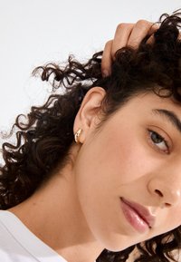 Guldøreringe med et glat, buet design. Øreringene præsenteres mod hud med krøllet hår delvist synligt.