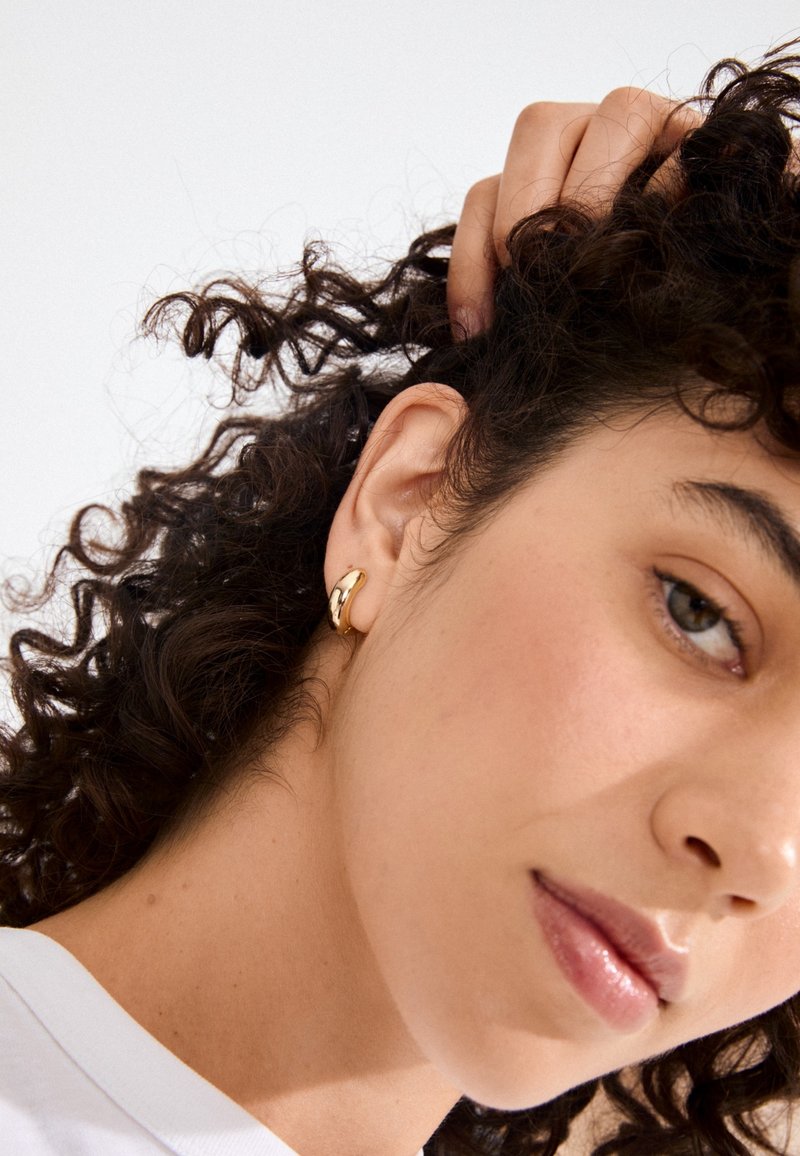 Guldøreringe med et glat, buet design. Øreringene præsenteres mod hud med krøllet hår delvist synligt.
