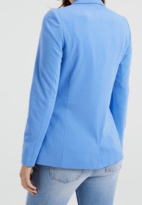 Modrý blejzr s přiléhavým střihem, se hladkým povrchem, dlouhými rukávy a švy na zadní straně. Nositý přes světle modré džíny.