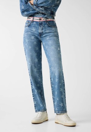 Osoba nosząca niebieskie dżinsy z wysokim stanem z wyblakłym wzorem gwiazd, biały pasek z czerwonymi detalami, białe trampki i kurtkę jeansową z wzorem gwiazd.