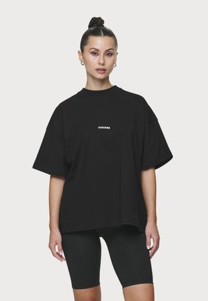 Sötét haját kontyba tűzött nő, arany karika fülbevalóval, túlméretezett fekete póló kis fehér felirattal, és fekete kerékpáros rövidnadrágban, semleges háttér előtt.