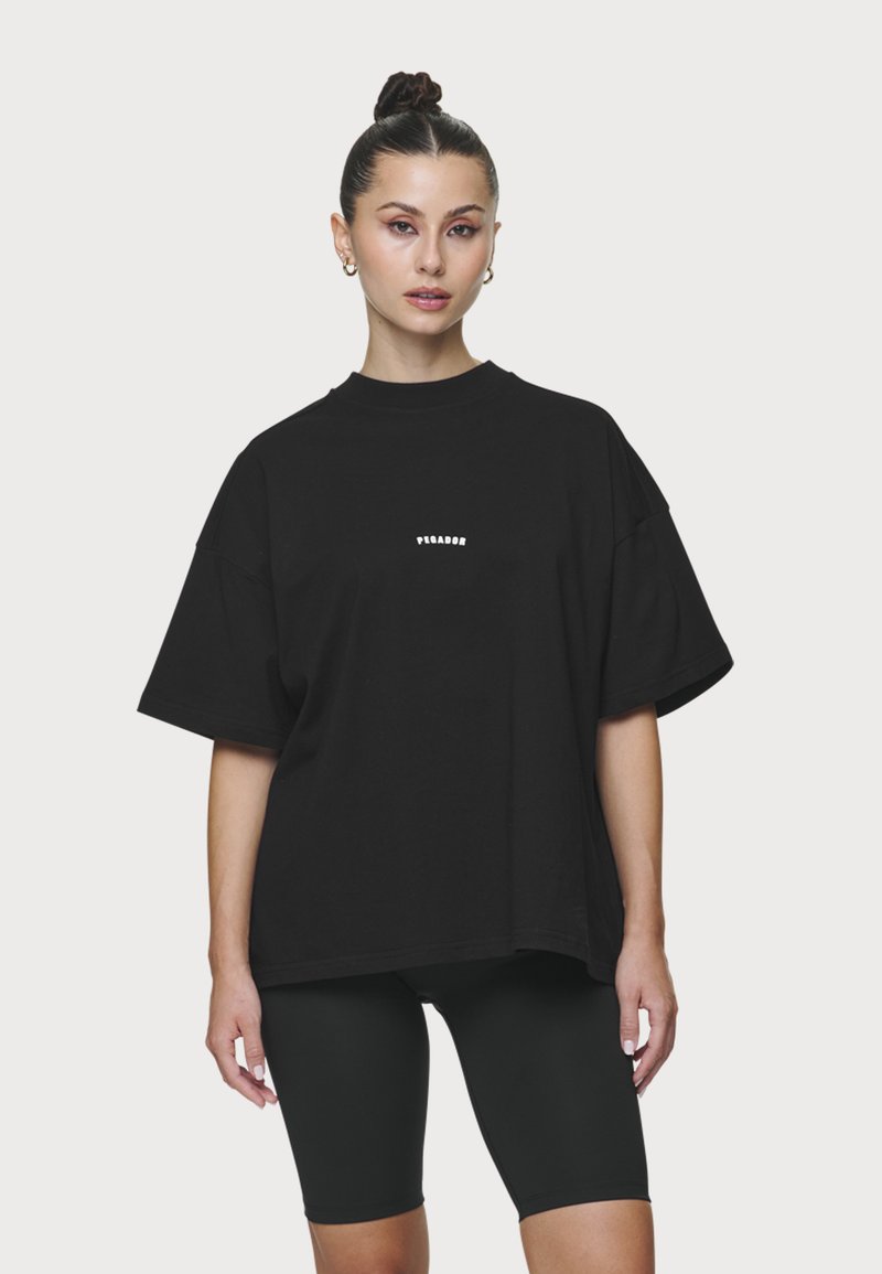 Γυναίκα με σκούρα μαλλιά δεμένα σε κότσο, φορά χρυσούς κρίκους στα αυτιά, υπερμεγέθη μαύρο T-shirt με μικρό λευκό κείμενο και μαύρα ποδηλατικά σορτς, σε ουδέτερο φόντο.