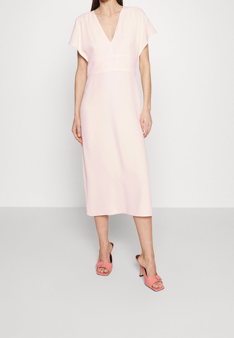Femme portant une robe midi rose pâle à manches courtes et encolure en V, associée à des sandales ouvertes à talons corail, debout sur un sol gris.