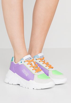 Benen met chunky sneakers met paarse, groene en witte panelen, oranje veters en "Versace Jeans Couture" branding aan de zijkant en tong.