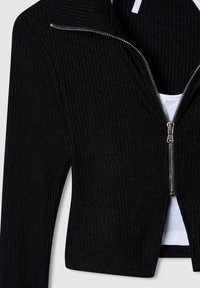 Giacca cropped nera a coste con chiusura frontale con zip, colletto alto e uno strato interno bianco liscio. Zip accentuata con dettagli metallici.