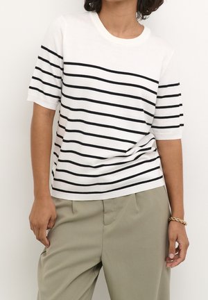 Personne portant une chemise blanche à manches courtes avec des rayures horizontales noires et un pantalon beige clair, debout devant un fond uni.