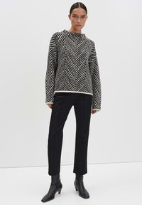 Frau in einem schwarz-weißen, im Zickzack-Muster gestrickten Pullover, schwarzen, eng anliegenden Hosen und schwarzen Ankle Boots, die vor einem einfarbigen Hintergrund steht.