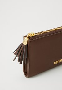 Portafoglio in pelle marrone con zip dorata e tirante a nappina, caratterizzato da testo parzialmente in rilievo dorato "LOVE MO" sulla parte anteriore.