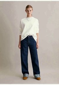 Mulher vestindo uma camisa branca de manga longa, calças largas escuras com bainhas, sapatos castanhos e um colar prateado, de pé contra um fundo plain.