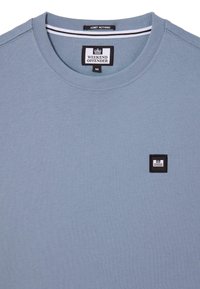 Tricou bleu deschis cu guler rotund, cu un mic patch cu logo-ul Weekend Offender în alb și negru pe pieptul stâng și eticheta "Admit Nothing" în interiorul gulerului.