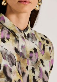 Close-up of a person wearing a patterned shirt with purple, black, and beige brushstroke designs and large gold hoop earrings.