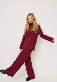 Bordeaux dobbeltbrystet blazer parret med matchende vide bukser. Har en glatt tekstur og gullfargede knapper, båret med svarte sko.