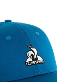 Gorra de béisbol azul de tela que presenta un logotipo bordado en blanco y negro de un gallo y el texto "LE COQ SPORTIF" en el frente.