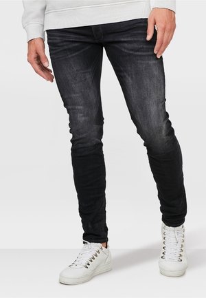 Man draagt slank gesneden zwart vervaagde jeans en witte sneakers, staand met ontspannen handen langs zijn zij tegen een effen achtergrond.