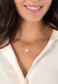 Zelta kaklarota ar teksturētu apaļu piekariņu, kurā iemontēts rozā akmens, valkāta virs baltās blūzes, izceļot smalkus ķēdes detaļas.