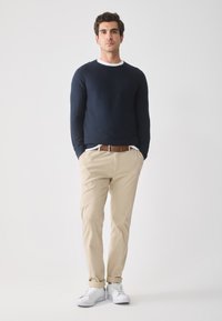Marine ribbelgebreide trui, kaki broek, bruine leren riem, witte shirtzoom zichtbaar en witte sneakers. Model staat tegen een lichte achtergrond.