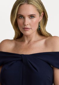 Jūras zils kleita ar atvērtām plecu daļām un plisēta priekšpuse. Modelis valkā zaļas auskarus. Gluda auduma sastāvs ar pieguļošu siluetu un dabiski spīguļojošu virsmu.