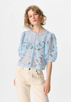 Donna con capelli biondi ricci che indossa una camicetta in pizzo leggero blu chiaro, trasparente, con bottoni e maniche a sbuffo, insieme a pantaloni beige a vita alta.