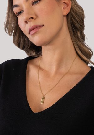 Young woman with light brown hair wears a black V-neck top and a gold rectangular pendant necklace engraved with "Be good".