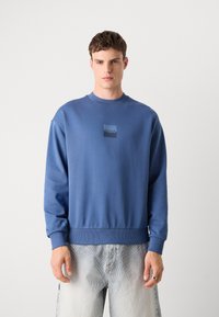 Modrá mikina vyrobená z mäkkého materiálu, s okrúhlym výstrihom, rebrovanými manžetami a spodným lemom, s štvorcovým logom "Hugo" na hrudi.