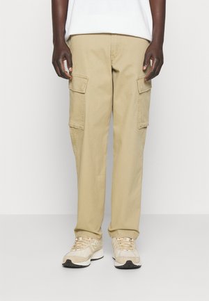 Persona che indossa pantaloni cargo beige con tasche a patta e sneaker beige New Balance, in piedi davanti a uno sfondo chiaro uniforme.