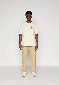 Κρεμ κοντομάνικο μπλουζάκι με λουλουδάτο λογότυπο, συνδυασμένο με μπεζ cargo παντελόνια. Το μοντέλο φοράει λευκά αθλητικά παπούτσια και ανοιχτόχρωμα γυαλιά ηλίου.