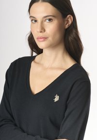 Donna con lunghi capelli scuri che indossa un maglione nero a V con un logo ricamato sul lato sinistro del petto, che guarda direttamente verso l'obiettivo.
