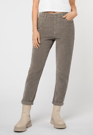 Persoon draagt opgerolde grijze corduroy broek, crèmekleurige stevige laarzen en een wit cropped top, van taille tot voeten tegen een effen achtergrond.