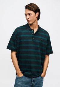 Jeune homme portant un polo à rayures horizontales bleu marine et vert avec une patte de boutonnage, les mains dans les poches, regardant sur le côté.