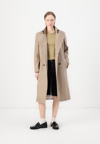 Beige trench coat with a double-breasted front, notch lapels, and long sleeves, worn over a green top and black shorts. Black shoes with white socks.