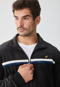 Zwarte zip-up jas met een zachte textuur, voorzien van blauwe en witte strepen op de borst en een klein geel logo op de bovenste linkerzijde.
