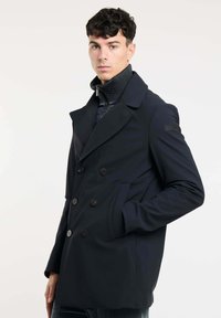 Marineblauwe, dubbelzijdige jas van een gladde stof met een geknipt kraag, grote knopen en zijzakken, gedragen over een binnenjas met hoge kraag.