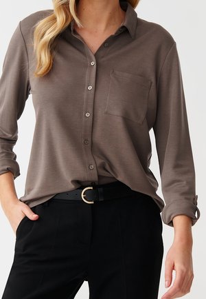 Frau trägt ein taupefarbenes Hemd mit Knopfleiste und Brusttasche, schwarzen Gürtel mit goldener Schnalle und schwarze Hose, eine Hand in der Tasche, Ärmel hochgekrempelt.