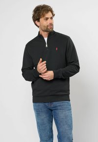 Crna zip-up dukserica s uspravnim ovratnikom i malim crvenim logom na lijevoj strani grudi. Mekani materijal, rebrasti manžeti i rub. Nosena s plavim trapericama.