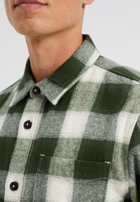 Green and white checkered flannel shirt with a collar, black buttons, and a front pocket. Soft texture with a warm and casual design.