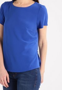 Krátký rukáv modrý top z hladké látky, s kulatým výstřihem, kapsou na prsou a bočním zipem. Nosit s tmavými džíny.