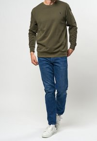 Oliivivihreä huppari pitkillä hihoilla ja pyöreällä kaula-aukolla, yhdistettynä sinisiin farkkuihin ja valkoisiin tennareihin, joissa on huomaamaton logoyksityiskohta.