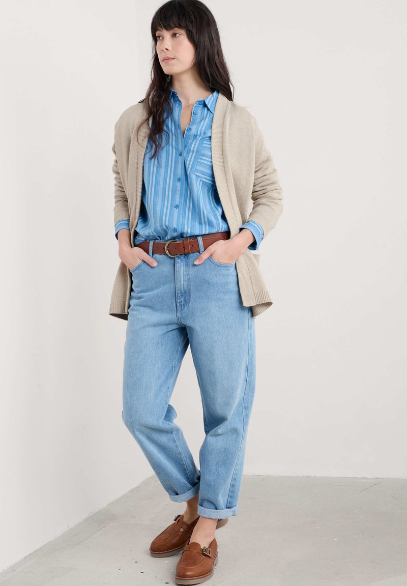 Bēšas adītas kardigāns pār zilu svītrainu krekli, augsto vidukļa gaismas zilo džinsu un brūnām ādas slip-on kurpēm; rokas kabatās.