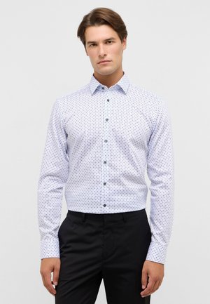 Witte button-up blouse met een marineblauw patroon van kleine stippen, lange mouwen en een standaard kraag; gecombineerd met zwarte broek.