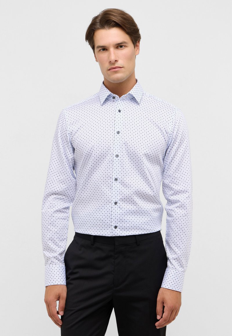 Camicia bianca con un motivo navy di piccoli punti, maniche lunghe e colletto standard; abbinata a pantaloni neri.