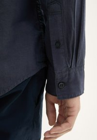 Primer plano de la mano de una persona descansando junto a la manga de su camisa de manga larga azul marino con dos botones negros y pantalones oscuros.