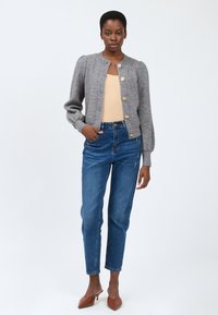 Cardigan grigio a maglia con bottoni dorati, abbinato a un top beige chiaro e jeans blu a vita alta. Il modello indossa mule con tacco marroni.