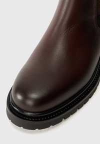 Bottes en cuir marron avec une finition lisse, bout rond et semelle en caoutchouc noir contrastante. Détails de couture le long du bord.