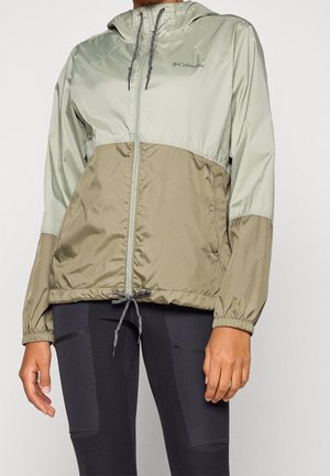Personne portant une veste coupe-vent Columbia bicolore vert clair et beige, avec un fermoir éclair frontal et un ourlet à cordon de serrage ajustable, accompagnée de leggings noirs.