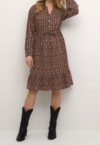 Vestido marrón con estampado, de manga larga y cierre de botones, con un dobladillo con volantes. Completan el look unas botas vaqueras negras.