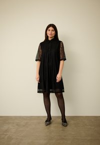 Melnā mežģīņu kleita ar īsām piedurknēm un pogu aizdari apkaklē. Brīvs piegriezums, ceļgalu garumā, kombinēta ar melnām zeķubikēm un apaviem zebras rakstā.