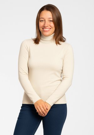 Maglione beige a collo alto con design aderente e maniche lunghe, realizzato in tessuto liscio. Abbinato a jeans blu scuro. La modella è in piedi con le mani incrociate.